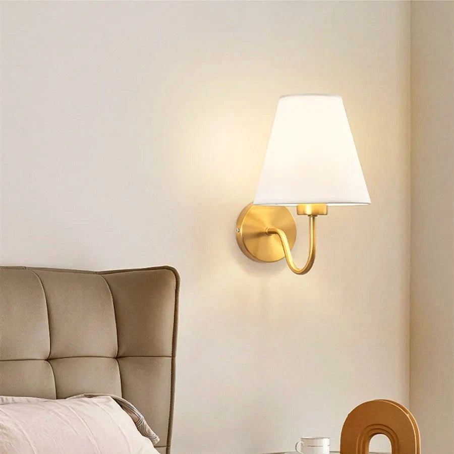 Brass wall sconce with off-white fabric shade mounted beside a bed