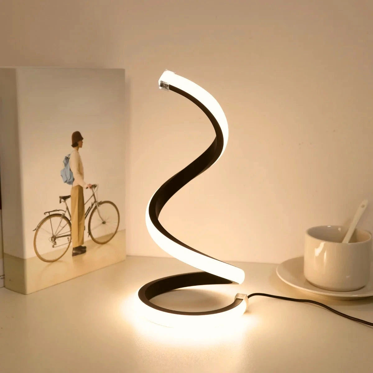Spiral LED table lamp glowing warm white on a desk beside a cup and book