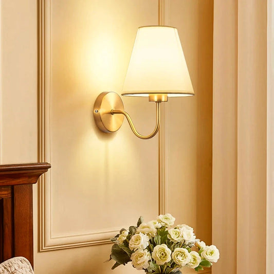 Linen shade wall sconce with curved brass arm lit above a bedside table